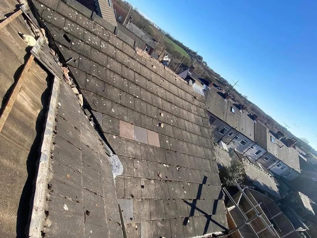 A worn tiled roof photographed from above, with cracked, slipped and mismatched patched tiles, looking out over a row of terraced houses.