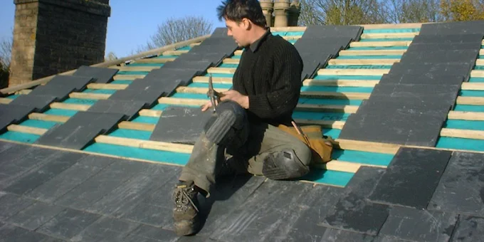 A roofer sitting on a roof, laying new dark slate over green waterproof lining and new timber battens.