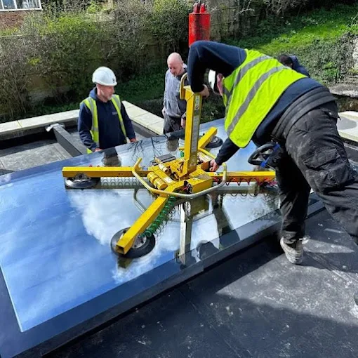 Four roofers in hi-vis lowering a large glass rooflight into a flat roof using a yellow vacuum lifter.