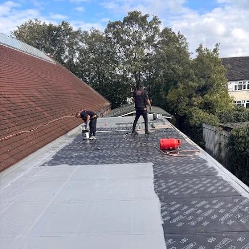 Two roofers working across a long flat roof, rolling out and sealing new layers of covering.